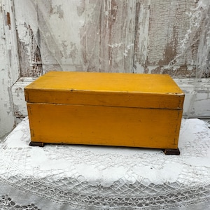 Puede incluir: Una pequeña caja de madera amarilla con tapa con bisagras y pequeños pies. La caja está sobre un mantel de encaje blanco.