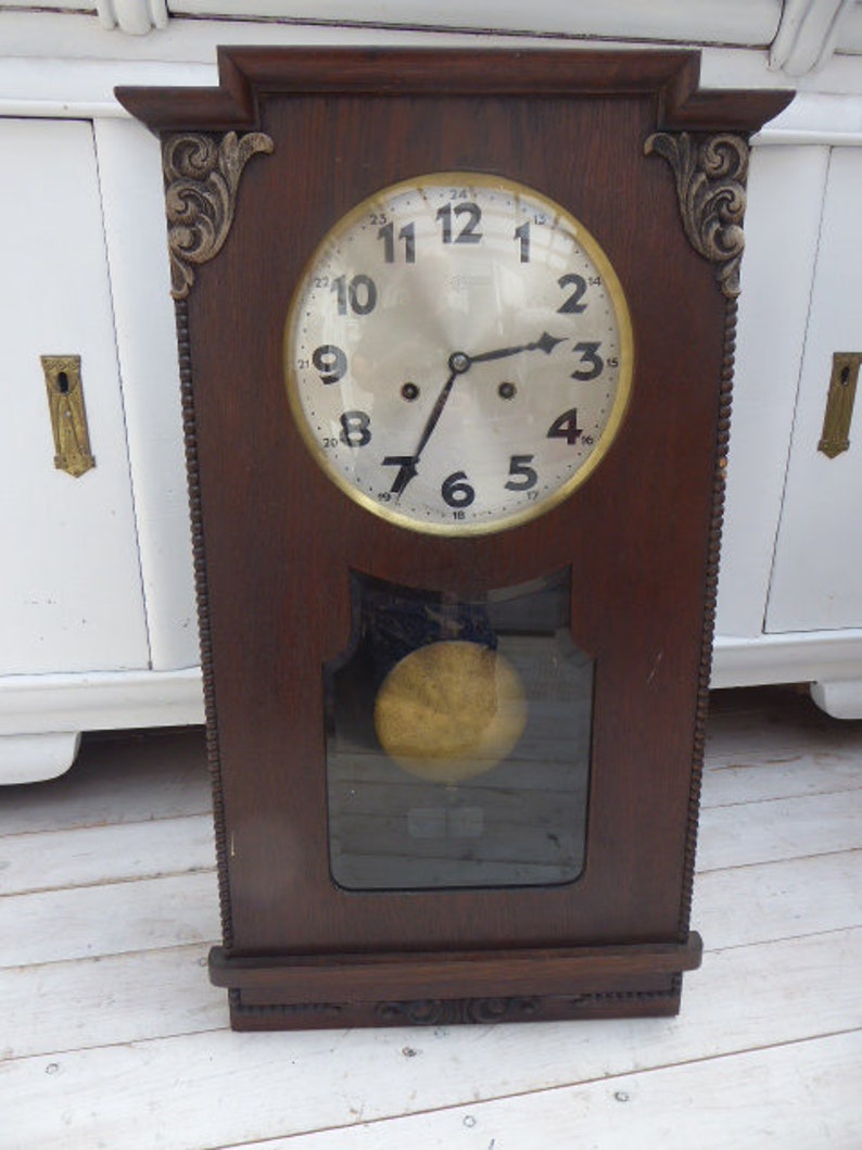 May include: Antique wooden wall clock with a round, white face and black Roman numeral markings. The clock has a decorative, ornate frame with gold accents.