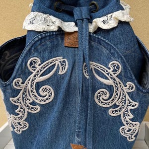 Op de afbeelding: Een blauwe denim rugzak met witte kantversiering en witte borduurwerk. De rugzak is 42 cm hoog, 28 cm breed en 17 cm diep.