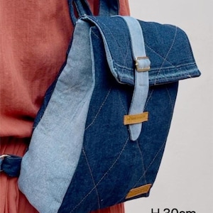May include: A denim backpack featuring a dark blue quilted main body with light blue side panels and a flap closure secured by a light blue strap and metal buckle. The bag measures 30 cm in height, 24 cm in width, and 10 cm in depth.