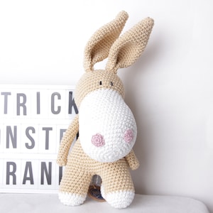 May include: A handmade, beige crocheted donkey plush toy with a white muzzle and pink accents. The donkey has long ears and is standing in front of a light box that reads "STRICK MONSTER BRAND".