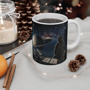Peut inclure: Un mug en céramique blanche orné d'un motif spatial sombre. Le design représente une personne portant un casque et un sac à dos, assise devant une grande planète avec des anneaux, une antenne parabolique et un carnet ouvert. Des constellations sont visibles.