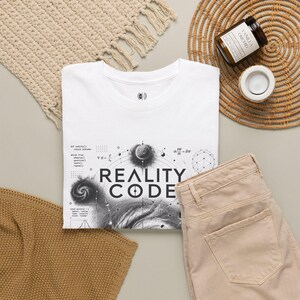 Op de afbeelding: Een wit t-shirt met een zwarte grafische print met de tekst "REALITY CODE" gecentreerd boven een wervelend nevelontwerp, wiskundige vergelijkingen en geometrische patronen. Het t-shirt heeft een ronde halslijn en een klein logo in de kraag.