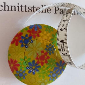 Könnte beinhalten: Ein weißes Maßband mit einem floralen Muster. Das Band ist in Zoll und Zentimetern gekennzeichnet. Das Etui ist gelb mit roten, blauen und grünen Blumen.