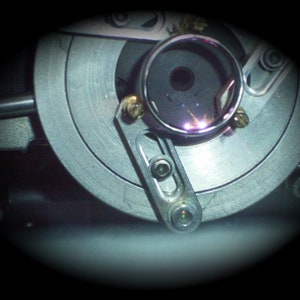 May include: Close-up of a mechanical device with a circular, metallic component. The device features a silver-colored, circular plate with a central opening and various screws and levers. A small, circular glass element is visible.