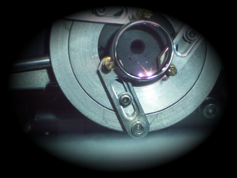 May include: Close-up of a mechanical device with a circular, metallic component. The device features a silver-colored, circular plate with a central opening and various screws and levers. A small, circular glass element is visible.