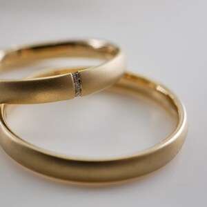 May include: Two gold wedding bands. One ring features a small section with embedded diamonds. The rings are a matte gold finish and are set against a white background. The rings are likely for a wedding or engagement.