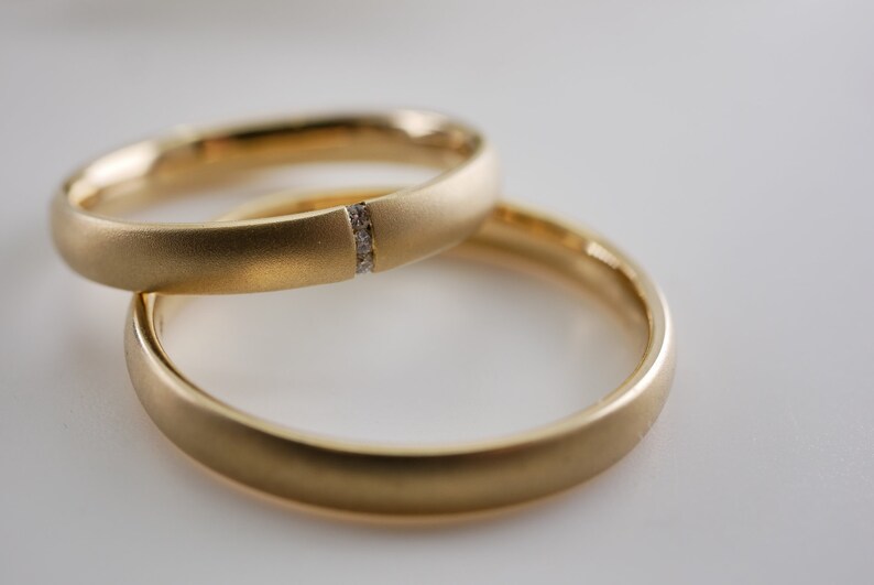 May include: Two gold wedding bands. One ring features a small section with embedded diamonds. The rings are a matte gold finish and are set against a white background. The rings are likely for a wedding or engagement.
