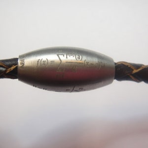 Könnte beinhalten: Eine silberne Metallperle mit einem geflochtenen Lederband. Die Perle ist mit einer mathematischen Formel graviert: "f(x) = Σ (f(0)(a) / (x - a)^n)".