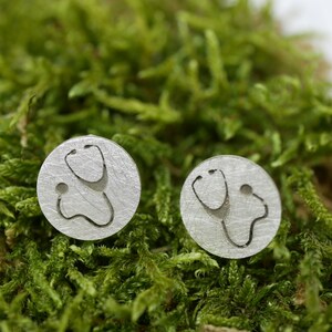 Könnte beinhalten: Ein Paar silberne Ohrringe mit einem Stethoskop-Design. Die Ohrringe sind rund und haben eine gebürstete Oberfläche.