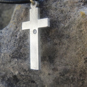 Könnte beinhalten: Ein silberner Kreuzanhänger mit einem kleinen, klaren Stein in der Mitte, der an einem schwarzen Lederband hängt.