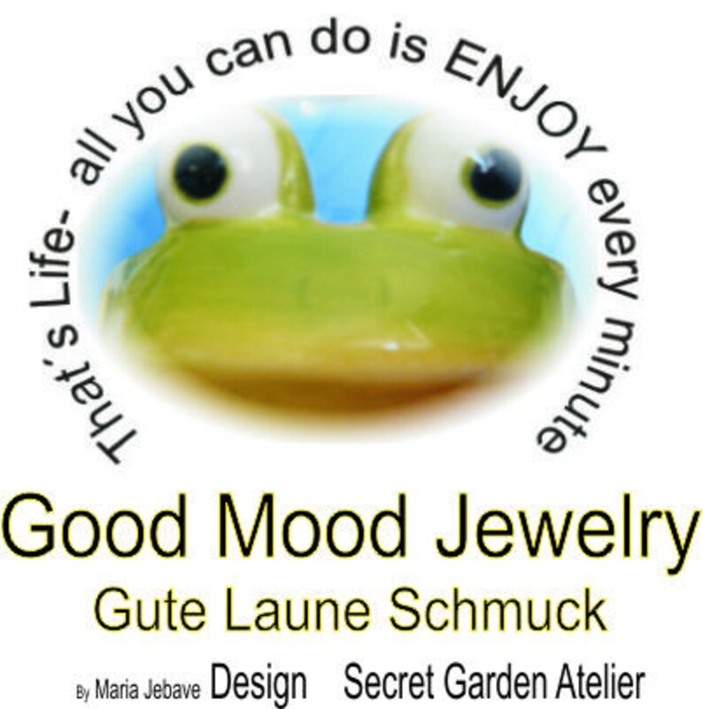 May include: A whimsical image featuring a green frog-like character with large, expressive eyes. The text "That's Life - all you can do is ENJOY every minute" arches around the frog. Below, the text reads "Good Mood Jewelry" and "Gute Laune Schmuck".