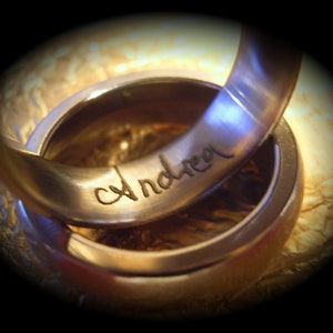 May include: Two intertwined gold wedding bands. The top band is engraved with the name "Andrea" in cursive script. The rings are set against a textured, golden background, with a dark vignette effect.