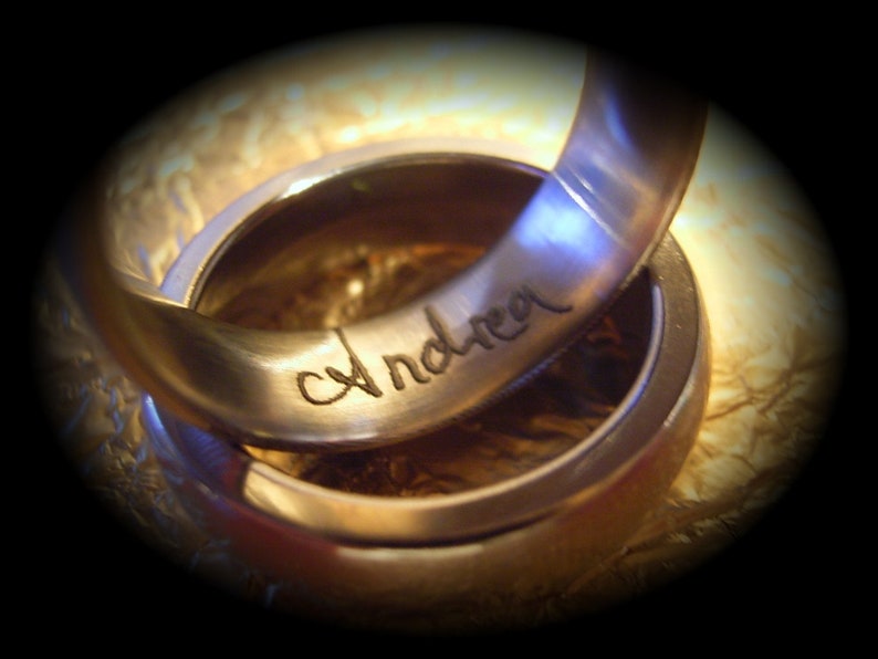 May include: Two intertwined gold wedding bands. The top band is engraved with the name "Andrea" in cursive script. The rings are set against a textured, golden background, with a dark vignette effect.