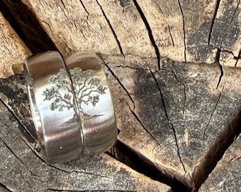 May include: Two silver rings with a tree design engraved on the surface. The rings are placed on a weathered, textured wooden surface. The tree design is detailed, with branches and leaves visible.