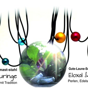 May include: A collection of necklaces with colorful, spherical beads in various hues, including blue, red, and green, suspended from black cords. The image includes text in German: "Damast-stahl Trauringe Unikate mit Tradition" and "Gute-Laune-Schmuck Eloxal & Co Perlen, Edelstahl & Natur."