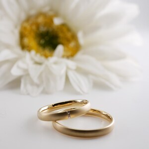 May include: Two gold wedding rings, one with a small diamond, are displayed in front of a blurred white daisy. The rings are smooth and have a matte finish, set against a bright white background.