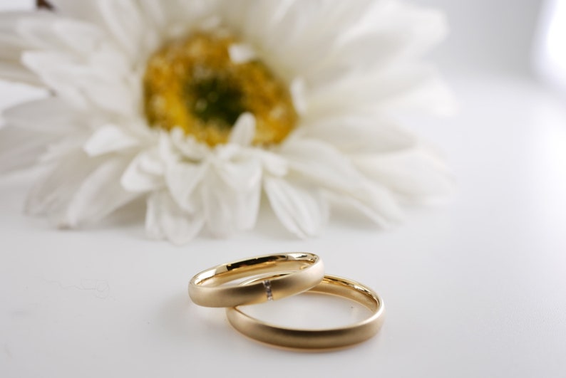 May include: Two gold wedding rings, one with a small diamond, are displayed in front of a blurred white daisy. The rings are smooth and have a matte finish, set against a bright white background.
