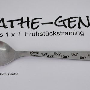 Könnte beinhalten: Ein silberner Löffel mit dem Text "MATHE-GENIE das 1 x 1 Frühstückstraining" und darauf gravierten Multiplikationsaufgaben.