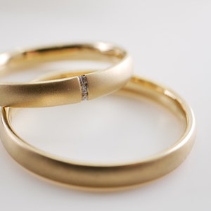 May include: Two gold wedding bands, one with a small diamond accent. The rings are a warm yellow color and have a matte finish. The bands are stacked on top of each other, with one slightly in front of the other.