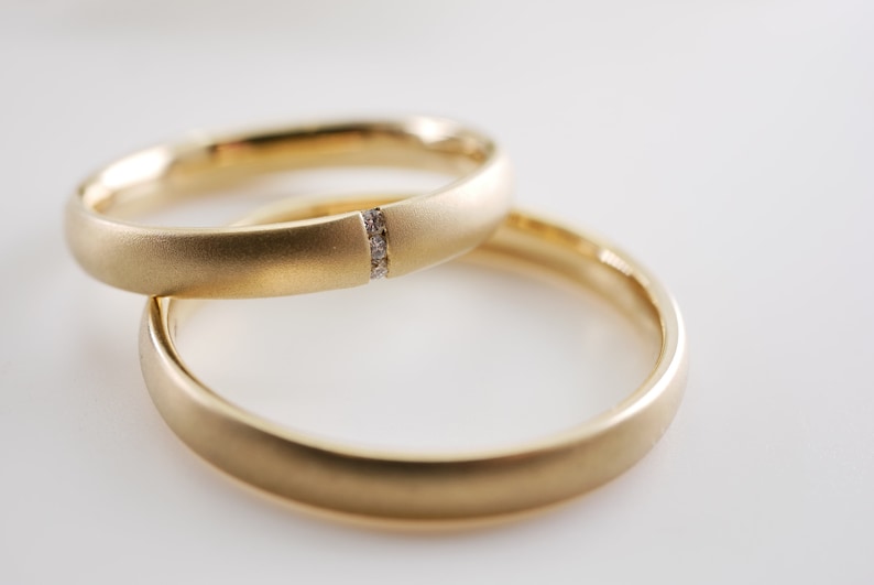 May include: Two gold wedding bands, one with a small diamond accent. The rings are a warm yellow color and have a matte finish. The bands are stacked on top of each other, with one slightly in front of the other.