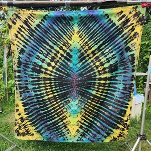 May include: A tie-dye textile with a radial pattern in black, yellow, blue, and purple hues. The fabric is displayed outdoors, suspended from a metal frame, showcasing its vibrant colors and unique design. The tie-dye pattern creates a symmetrical, eye-catching visual.