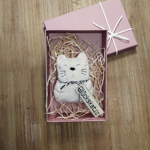 Könnte beinhalten: Ein weißes Plüschkatzenspielzeug mit schwarzen gestickten Augen und Schnurrhaaren, das in einer rosa Box mit zerkleinertem Papier sitzt. Das Katzenspielzeug hat ein Etikett mit der Aufschrift "Glückskatze".