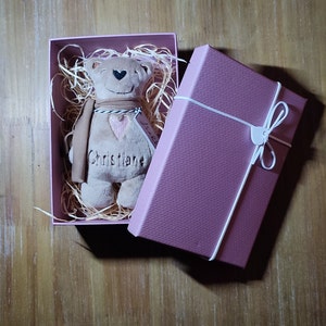 Könnte beinhalten: Ein plüschiger brauner Teddybär mit einem rosa Herzen und dem Namen "Christiane" auf der Brust gestickt, der in einer rosa Geschenkbox mit zerknülltem Papier sitzt.