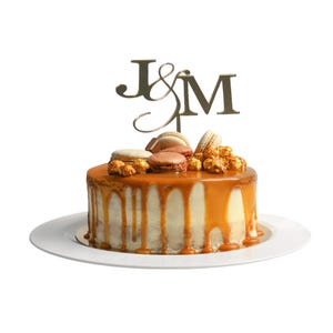 May include: A white cake with caramel frosting and a gold topper that says "J&M". The cake is decorated with caramel popcorn and macarons.