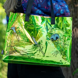 Op de afbeelding: Een glanzende groene draagtas met zwarte handvatten. De tas heeft cartoon illustraties van een schedel, botten en zwaarden. De tas wordt vastgehouden door een persoon die een blauwe top met bloemen draagt.