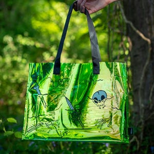 May include: A green iridescent tote bag with black handles, featuring a skull and crossbones design, along with swords and bone graphics. The bag is held up against a backdrop of green foliage and blue flowers.