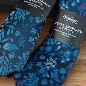 Puede incluir: Dos corbatas azul marino inspiradas en TTRPG con un patrón floral y geométrico en tonos azules y morados. Las corbatas están etiquetadas como "WitchHunt TTRPG-INSPIRED NECKTIE" y están hechas a mano con 100% poliéster.