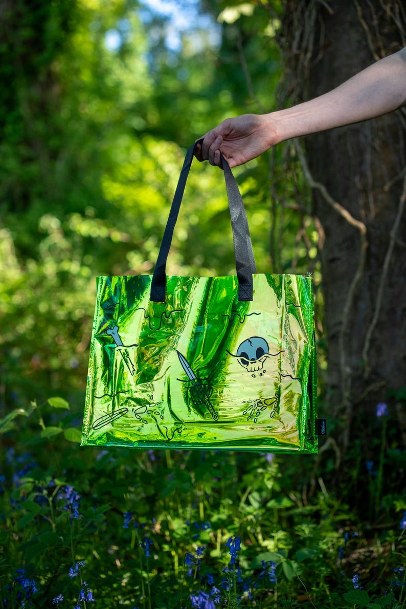 May include: A green iridescent tote bag with black handles, featuring a skull and crossbones design, along with swords and bone graphics. The bag is held up against a backdrop of green foliage and blue flowers.