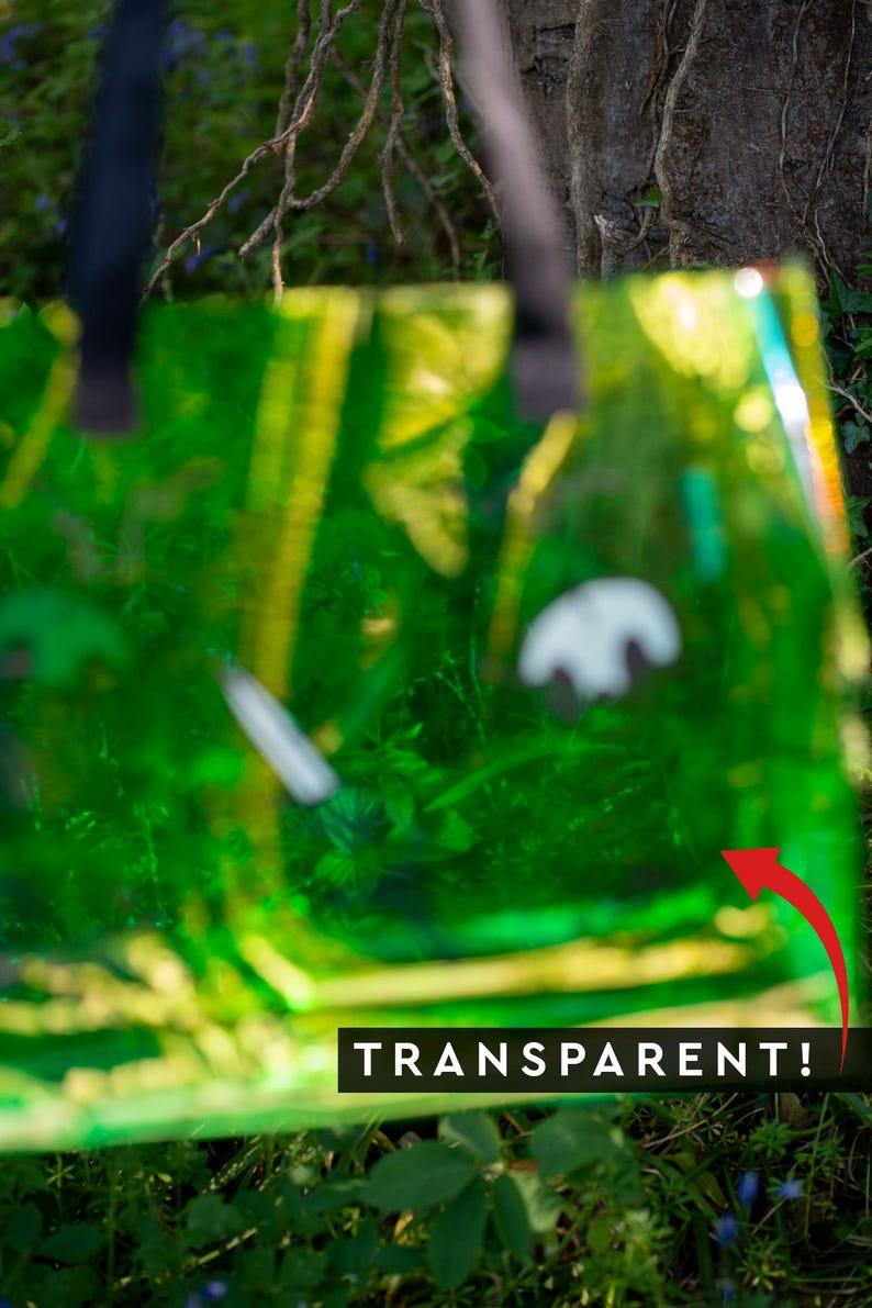 May include: A transparent green tote bag with black handles. The bag is filled with green foliage and has the word "TRANSPARENT!" in a white font. The bag is outdoors, with a blurred background of trees and plants.
