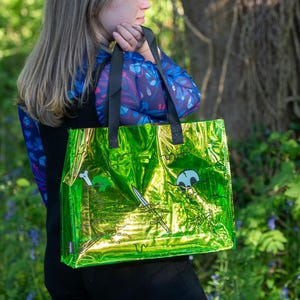 May include: A shiny green tote bag with black handles. The bag features a pirate-themed design with a skull, crossed swords, and a bone. The bag is held by a person in a blue floral patterned top.