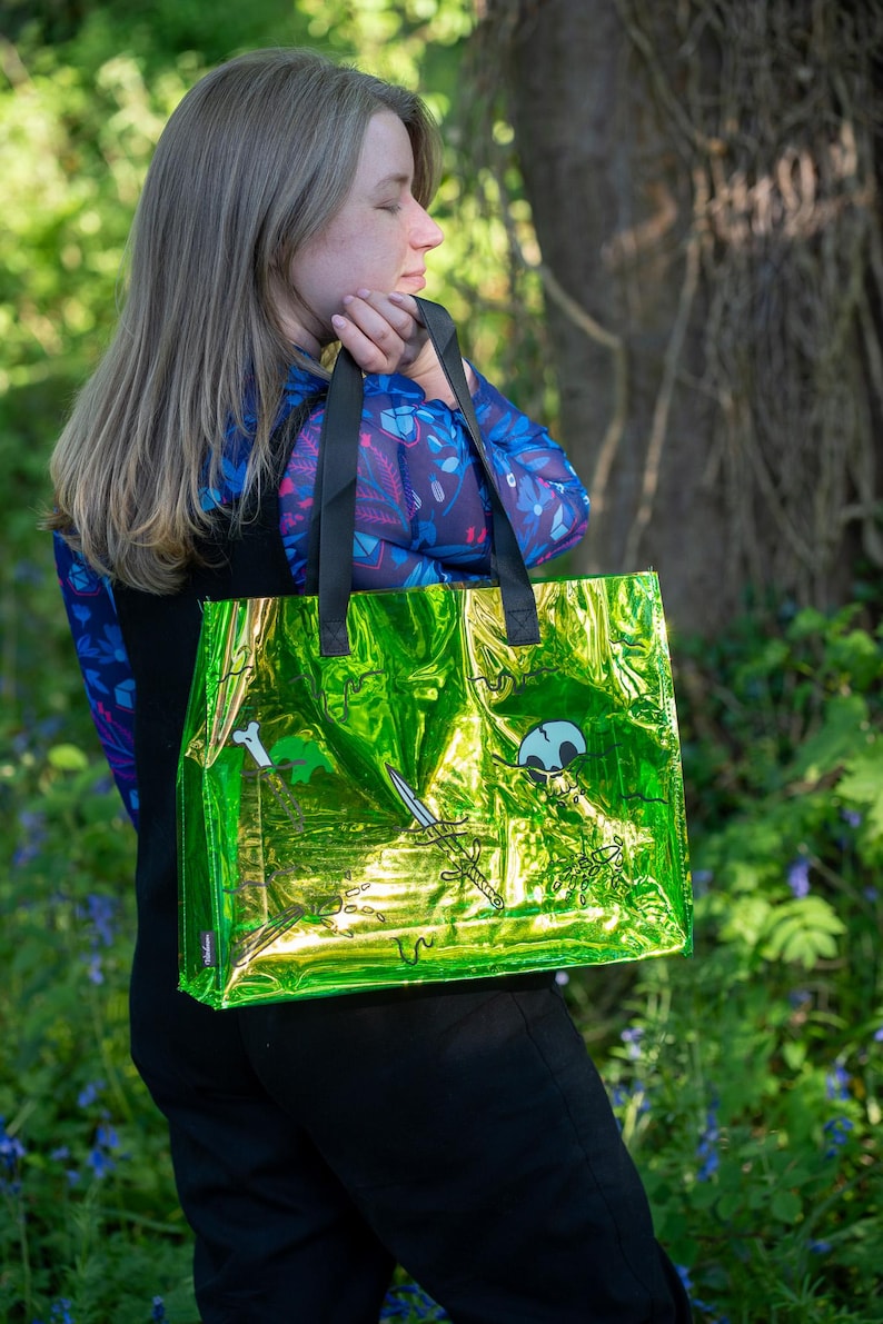 May include: A shiny green tote bag with black handles. The bag features a pirate-themed design with a skull, crossed swords, and a bone. The bag is held by a person in a blue floral patterned top.
