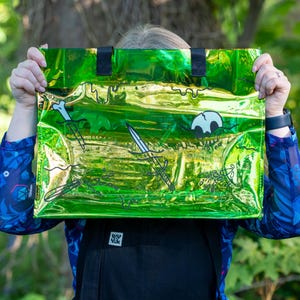 May include: A green and gold tote bag with black handles. The bag features a design of a skull, swords, and bones. The bag is held up in front of a person.