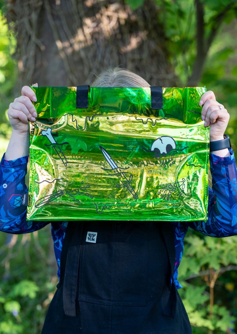 May include: A green and gold tote bag with black handles. The bag features a design of a skull, swords, and bones. The bag is held up in front of a person.