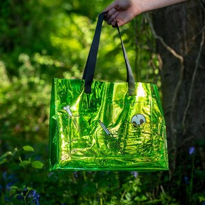 May include: A bright green tote bag with black handles, held against a blurred forest backdrop. The bag has a shiny, metallic finish and features a black and white illustration of a skull, sword, and bone. The bag is a reusable shopping bag.