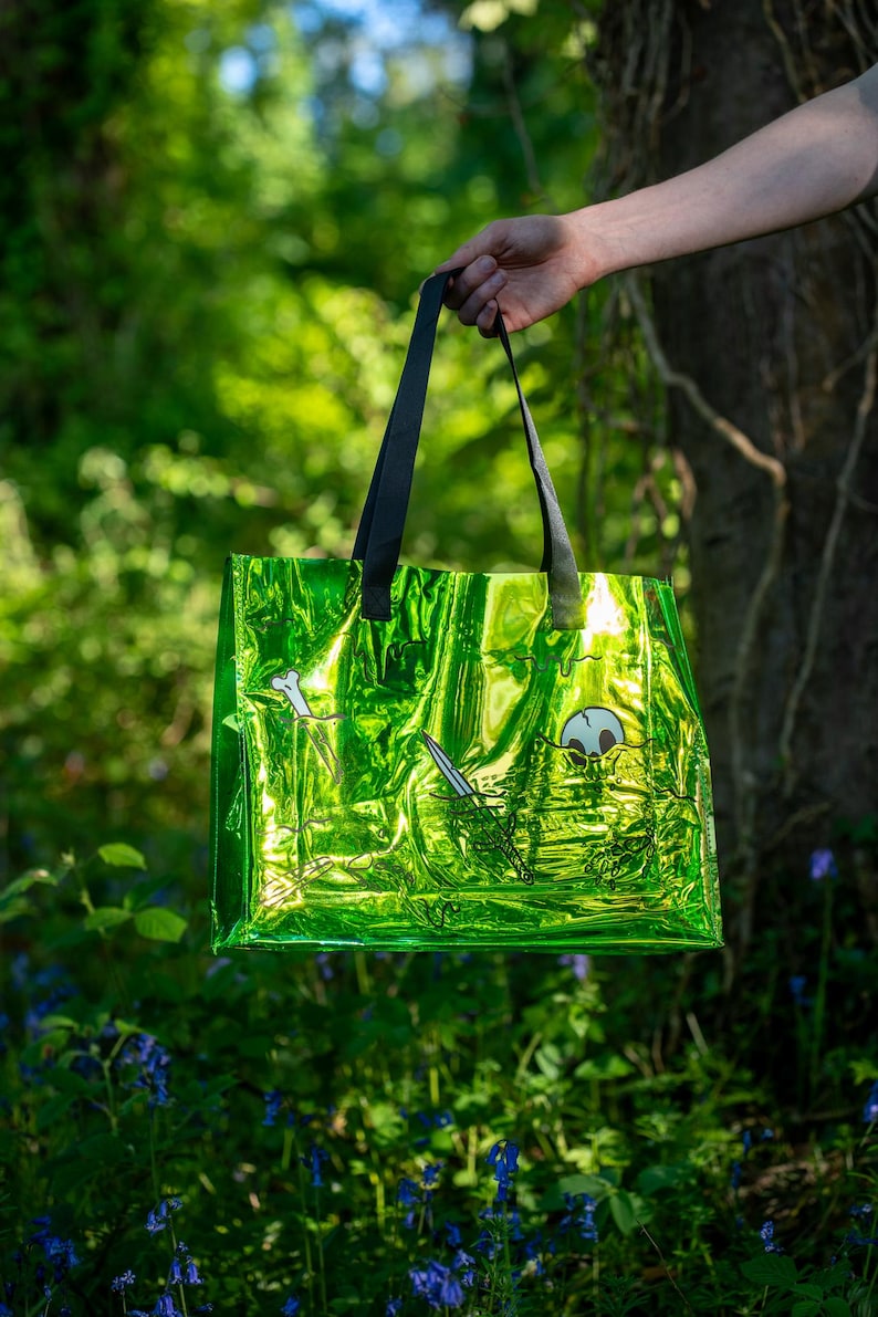 May include: A bright green tote bag with black handles, held against a blurred forest backdrop. The bag has a shiny, metallic finish and features a black and white illustration of a skull, sword, and bone. The bag is a reusable shopping bag.
