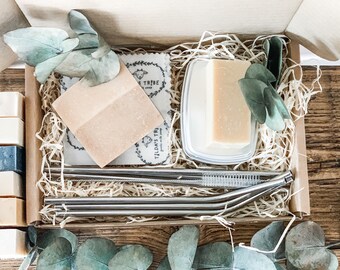 May include: A gift box filled with eco-friendly products, including two bars of soap, a soap dish, a set of stainless steel straws, and a reusable cloth. The box is lined with shredded paper and decorated with eucalyptus leaves.
