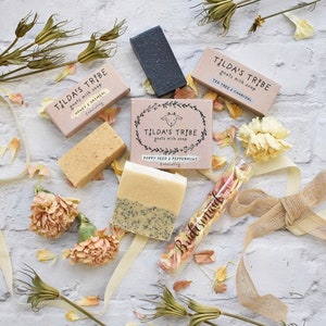 May include: A set of four goat milk soap bars with different scents and ingredients. The soaps are packaged in brown boxes with white text and illustrations. The boxes are arranged on a white surface with dried flowers and a small glass vial filled with flower petals.