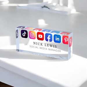 May include: A clear glass desk plaque with the text "NICK LEWIS SOCIAL MEDIA MANAGER" and icons for TikTok, Instagram, YouTube, Facebook, LinkedIn, and Pinterest.