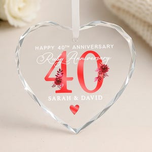 May include: A clear, heart-shaped crystal ornament with a white ribbon. It features the text "HAPPY 40TH ANNIVERSARY" and "Ruby Anniversary". The number "40" is red with floral details, and the names "SARAH & DAVID" are below. Ideal for a 40th wedding anniversary.