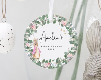 May include: A round, white ornament with a green, floral wreath design. The wreath features a small, pink-clad bunny, pink flowers, and green leaves. The text "Amelia's First Easter 2023" is printed in black on the ornament.