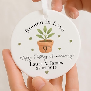 May include: A white ceramic round ornament with the text "Rooted in Love" and "Happy Pottery Anniversary". It features a potted plant graphic and the names "Laura & James" with the date 28.09.2016. A white ribbon is attached for hanging.