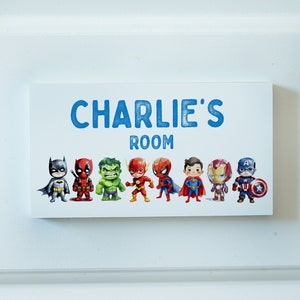 May include: A white wooden door sign with the text "CHARLIE'S ROOM" in blue lettering. The sign features a row of eight cartoon superheroes, including Batman, Deadpool, Hulk, Flash, Spiderman, Superman, Iron Man, and Captain America.