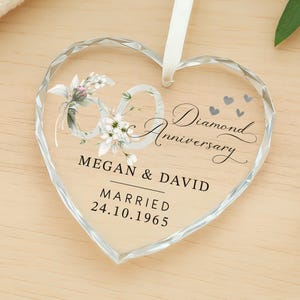 May include: A clear, heart-shaped crystal ornament with a white ribbon. It displays the text "60 Diamond Anniversary", the names "Megan & David", and the date "24.10.1965". The design is embellished with floral details.