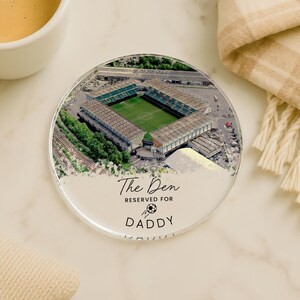 May include: A round coaster with an aerial view of a stadium, featuring a green field and seating. The coaster has the text "The Den RESERVED FOR DADDY" with a soccer ball graphic. The coaster is on a marble surface.