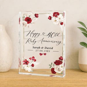 May include: A clear acrylic block with a floral design and the text "Happy 40th Ruby Anniversary." The design includes red and white flowers, with the names "Sarah & David" and the date "27.08.1985" below the text.